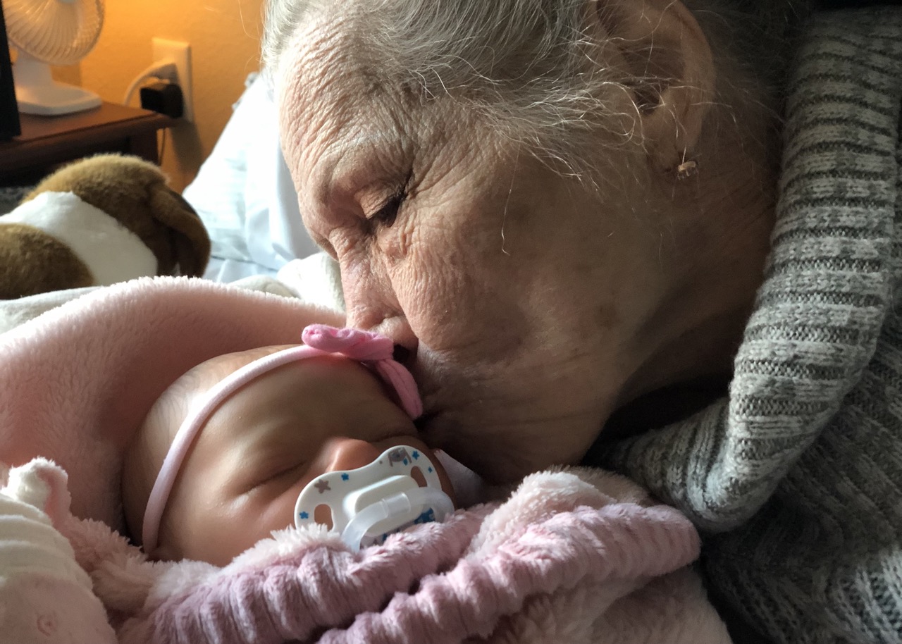 An elderly woman kisses a reborn therapy doll, wrapped in a pink blanket. The baby is wearing a pink bow headband and has a pacifier attached. The elderly woman is wearing a green sweater and is sitting on a bed.